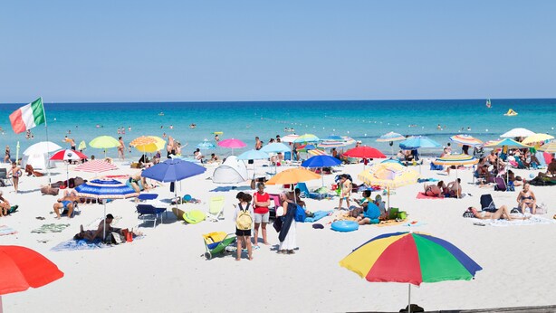 Een druk strand met mensen die zonnebaden en zwemmen, kleurrijke parasols, en een helderblauwe lucht.