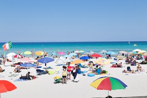 Une plage bondée où les gens se prélassent au soleil et se baignent, des parasols aux couleurs vives et un ciel bleu dégagé.