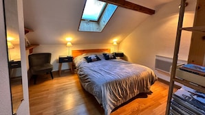 Quarto com clarabóia, soalho de madeira, cama com almofadas, duas mesas de cabeceira com luminárias e uma cadeira.
