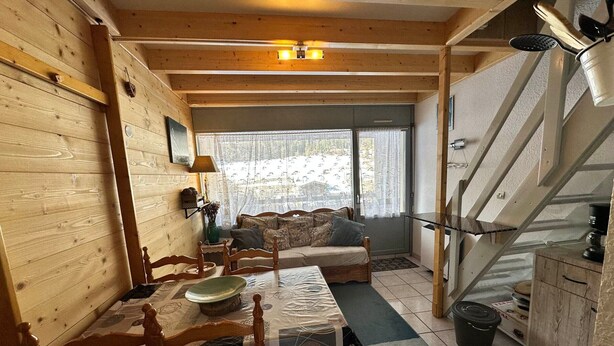 Interior acolhedor de cabana com mesa de jantar de madeira, cadeiras, sofá, escada e área de cozinha.