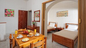A room with a wooden dining table set with plates, a bowl of fruit, and a glass of orange juice. There are two beds with wooden frames and white bedding. The walls are white with floral paintings.
