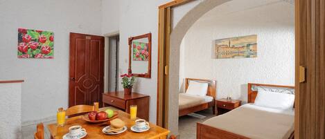 Una habitación con una mesa de comedor de madera dispuesta con platos, un cuenco de fruta y un vaso de zumo de naranja. Hay dos camas con estructura de madera y ropa de cama blanca. Las paredes son blancas y decoradas con pinturas florales.