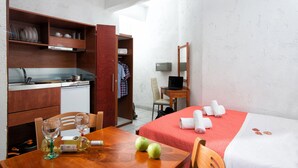 A compact hotel room with a kitchenette, a dining area, a bed with a red bedspread, and a mirror. - Cozy Studio in Hersonissos Center (Hersonissos)