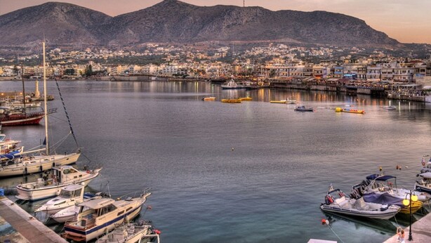 A marina with boats docked, a calm sea, and a cityscape with buildings and a mountain in the background. - Cozy Studio in Hersonissos Center (Hersonissos)