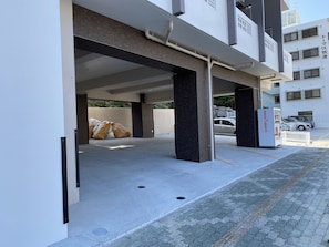 A parking garage with concrete floors and pillars, a vending machine, and a building with multiple windows and balconies in the background. - Hotel Tomas Makiminato Villa (Urasoe)