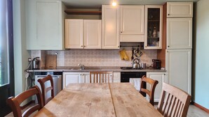 Cocina con armarios blancos, una mesa de madera y sillas. Se observa una ventana a la izquierda, y una lámpara colgante.