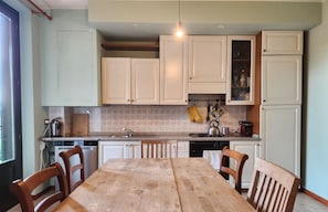 Una cucina con mobili bianchi, un tavolo di legno e sedie. Si vede una finestra a sinistra e un lampadario.