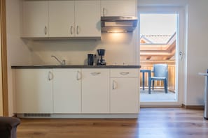 Une cuisine avec des meubles blancs, une machine à café et une fenêtre donnant sur un espace extérieur aménagé pour s’asseoir.