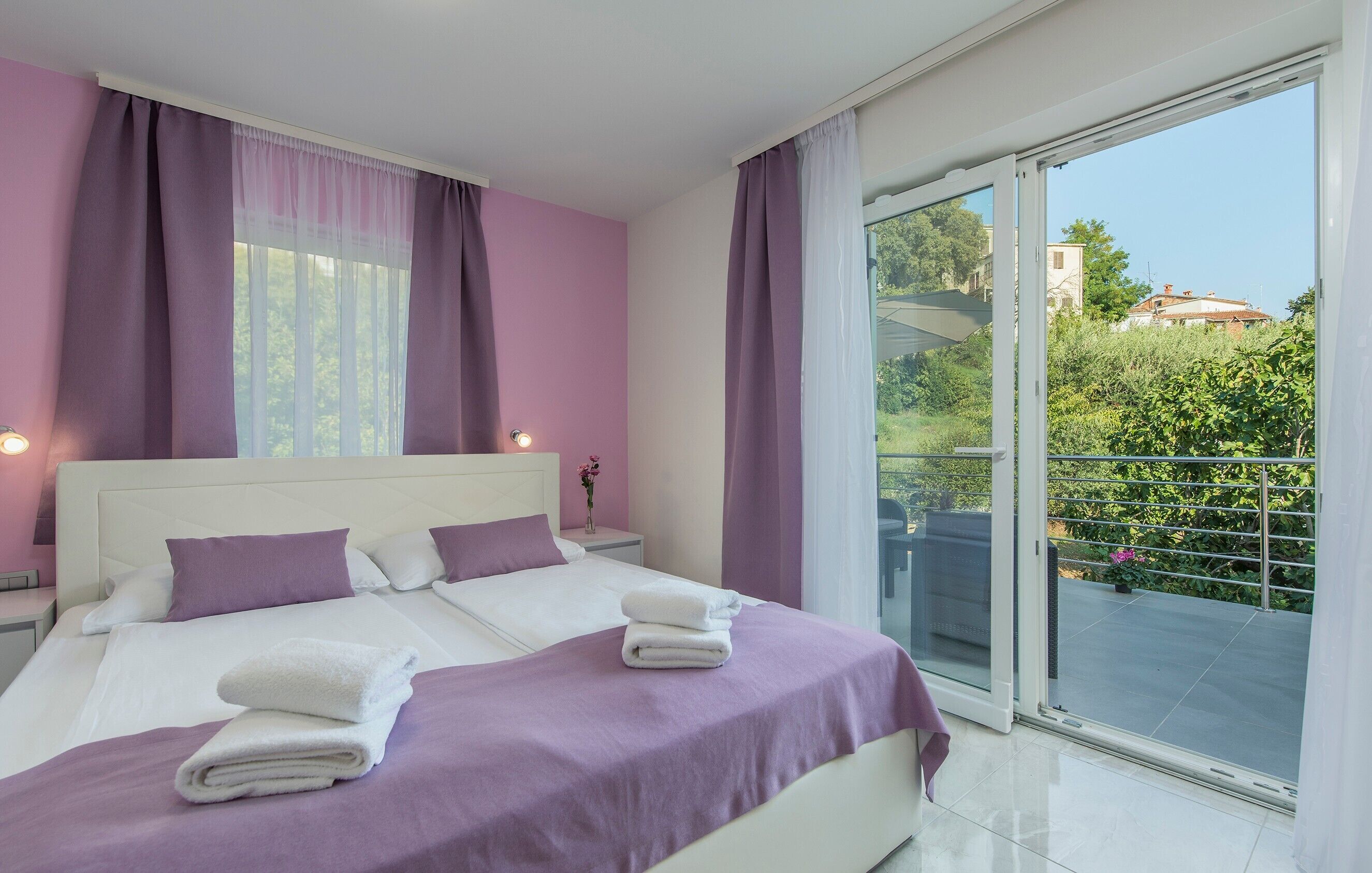 A bedroom with a large bed, two bedside tables, a nightstand, and a view of a balcony with greenery.