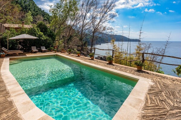 A rectangular pool with clear water, surrounded by lounge chairs and potted plants, overlooking a body of water and hills.