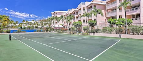 A tennis court with a net, surrounded by a fence and palm trees, with a building in the background.