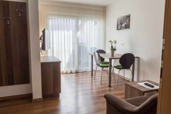 Un salon moderne avec un plancher en bois, un canapé, une table à manger avec des chaises, un téléviseur et une fenêtre ornée de rideaux légers.