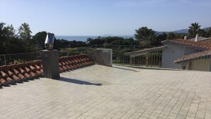 Una terraza en la azotea con suelo de baldosas, barandilla metálica y vistas al mar y las colinas.