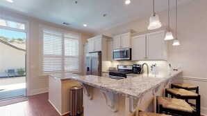 A kitchen with white cabinets, a granite countertop, a microwave, a refrigerator, and pendant lights. - Ocean Cottage 114 The Cottages at Jekyll Island Oceanfront (Jekyll Island)