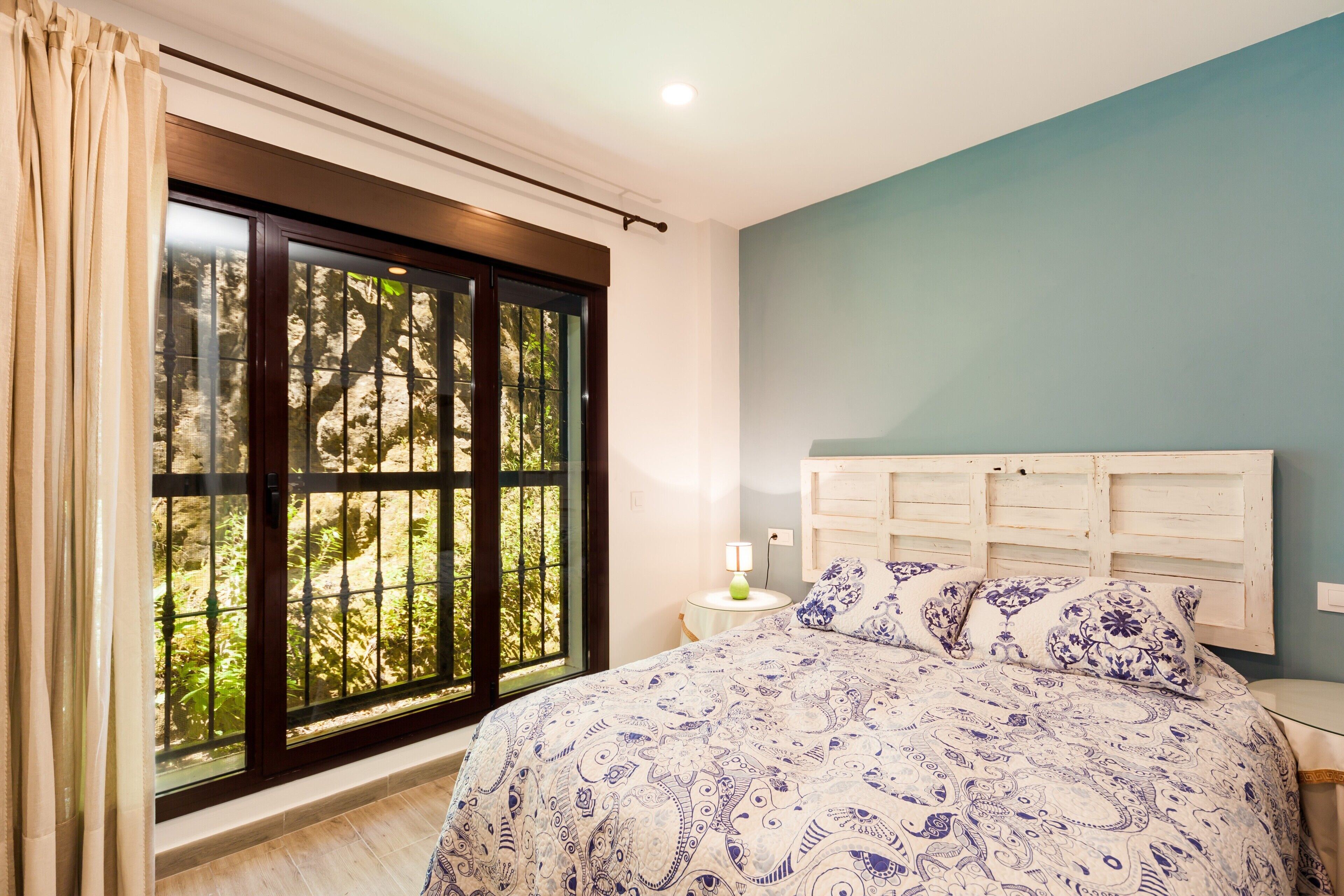 Ein Schlafzimmer mit großem Fenster, einem Bett mit gemusterten Betttextilien, einem Nachttisch mit Lampe und einem Holz-Kopfteil.