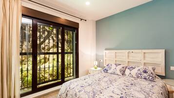 Ein Schlafzimmer mit großem Fenster, einem Bett mit gemusterten Betttextilien, einem Nachttisch mit Lampe und einem Holz-Kopfteil.