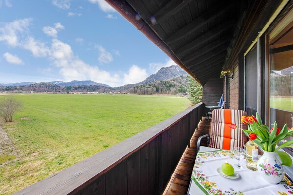 A balcony with a view of a green field and mountains, a table with a vase of flowers, and a chair with striped cushions. - Apartment 1 with Mountain View, Wi-Fi & Balcony (Grassau)