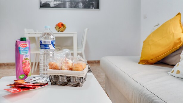 Une pièce avec un canapé blanc, un coussin jaune, un panier en osier contenant du pain, une bouteille d’eau, un emballage de jus de fruits et un tableau encadré accroché au mur.