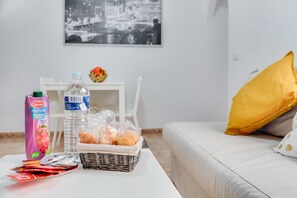 A room with a white sofa, a yellow pillow, a wicker basket with bread, a water bottle, a carton of juice, and a framed picture on the wall.