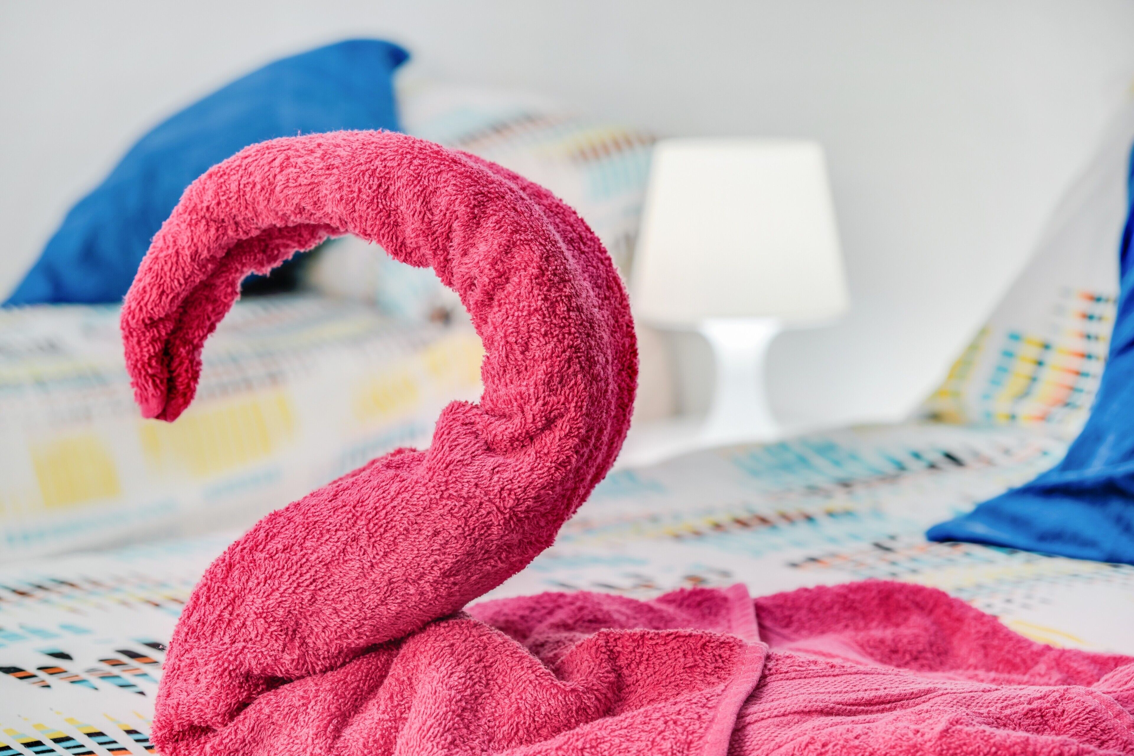 Ein Bett mit einem pinkfarbenen Handtuch, geformt wie ein Schwan, einer weißen Lampe und einem blauen Kopfkissen in einem Hotelzimmer.