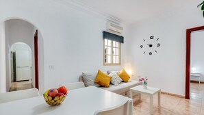 Un salon moderne avec un canapé blanc, une table à manger, un panier de fruits, une horloge murale et une fenêtre avec des stores.