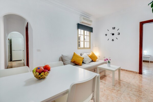 A modern living room with a white sofa, a dining table, a fruit bowl, a wall clock, and a window with blinds.