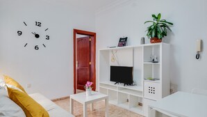 Une pièce avec un canapé blanc, une petite table blanche, un téléviseur posé sur un meuble blanc, une plante en pot et une horloge murale.