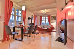 Ein gemütliches Wohnzimmer mit einem roten Sofa, einem Essbereich mit Tisch und Stühlen sowie einem an der Wand befestigten Fernseher.