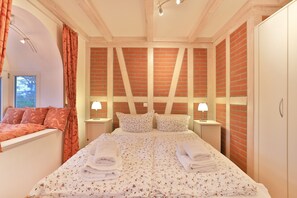 A bedroom with a large bed, two bedside tables with lamps, a window with curtains, and a brick-patterned wall.