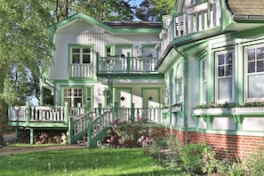 A two-story house with a green and white exterior, a front porch with a staircase, and a brick foundation.