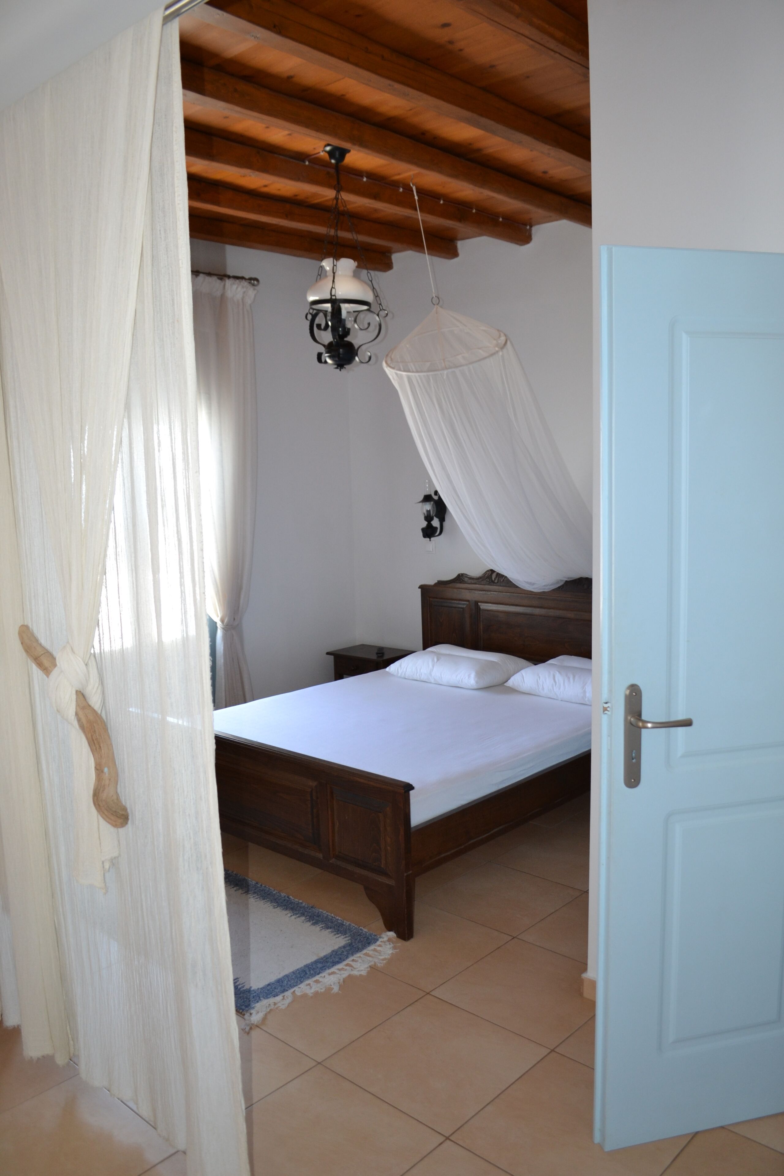 Un dormitorio con techo de madera, una cama con ropa blanca, una mesita de noche, una lámpara colgante y una puerta.