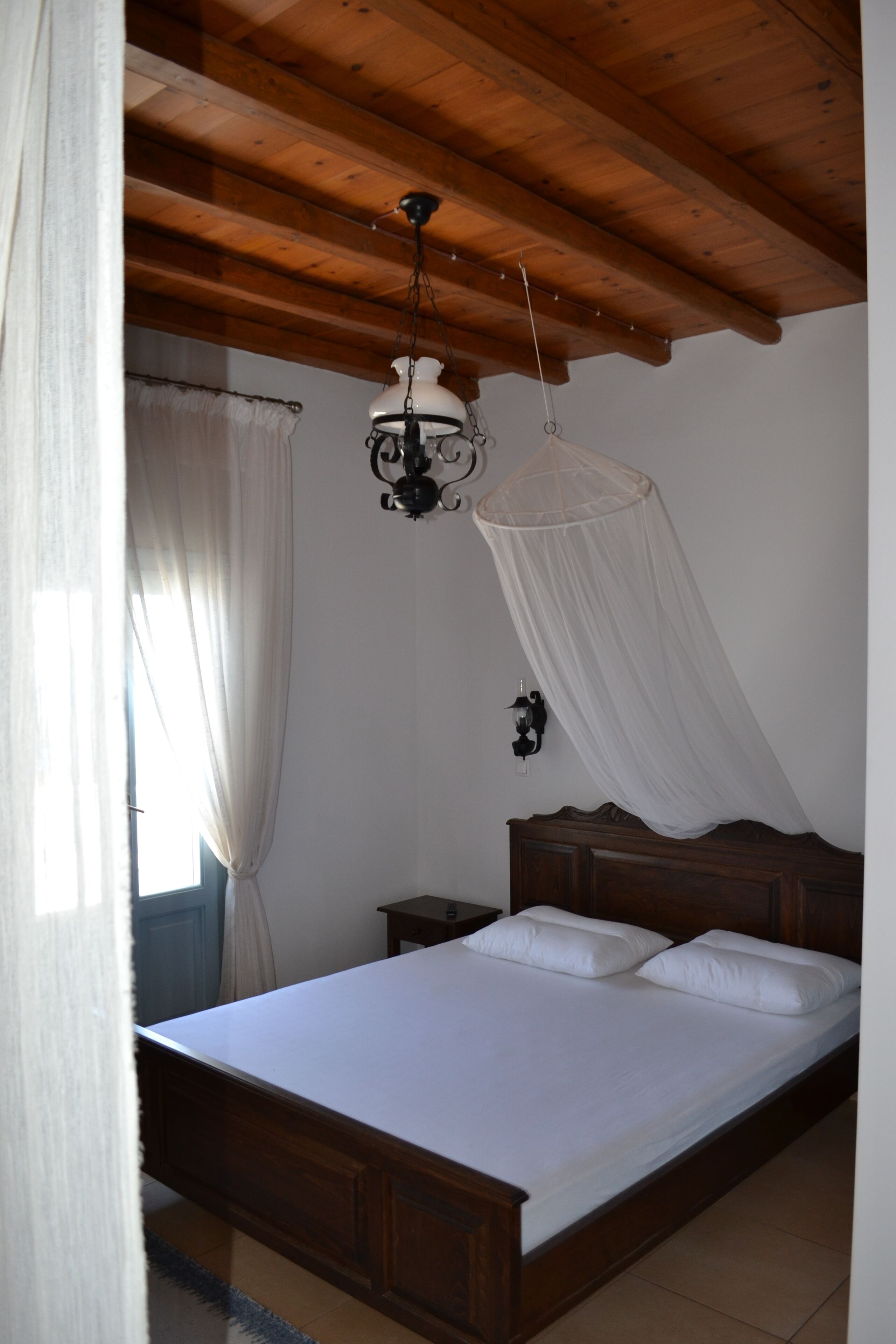 A bedroom with a wooden ceiling, a bed with white bedding, a hanging light fixture, and a nightstand.