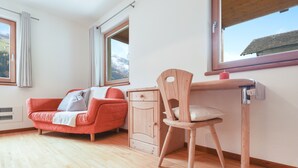 Une pièce avec un plancher en bois, un canapé rouge, une chaise en bois, un bureau et une fenêtre donnant sur les montagnes.