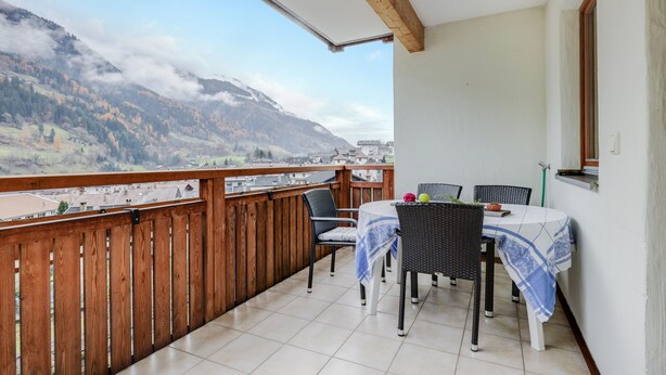Ein Balkon mit Holzgeländer, einem Tisch und Stühlen sowie Blick auf Berge und eine Stadt.