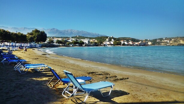 Une plage bordée de rangées de chaises longues bleues, un rivage sablonneux et des bâtiments en arrière-plan.