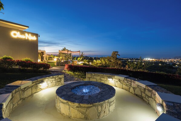 Un foyer en pierre donnant sur une vue panoramique de la ville au crépuscule, un bâtiment avec une enseigne lumineuse indiquant « Olive Nest », et des jardins aménagés.