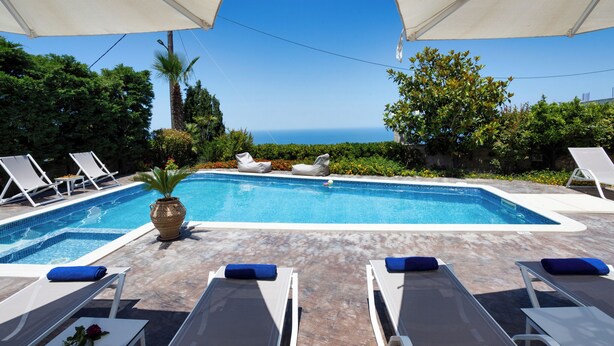 A swimming pool with lounge chairs and umbrellas, surrounded by greenery and a view of the ocean. - Palladio Luxury Villa With Amazing View (Rethymno)