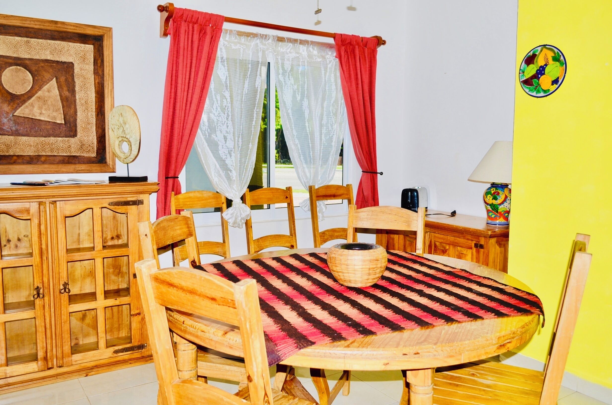 Un comedor con una mesa redonda de madera, mantel a rayas rojas y negras, y una cesta tejida. Hay sillas de madera alrededor de la mesa, y se puede ver un mueble de madera con puertas de vidrio. Cortinas rojas cubren las ventanas, y un cuadro enmarcado cuelga en la pared.