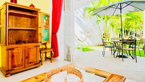 A room with a wooden cabinet, a wicker basket, a dining table with chairs, and a view of a garden with palm trees. - Room-playacar - Superior Budget Room Wit (Playa del Carmen)