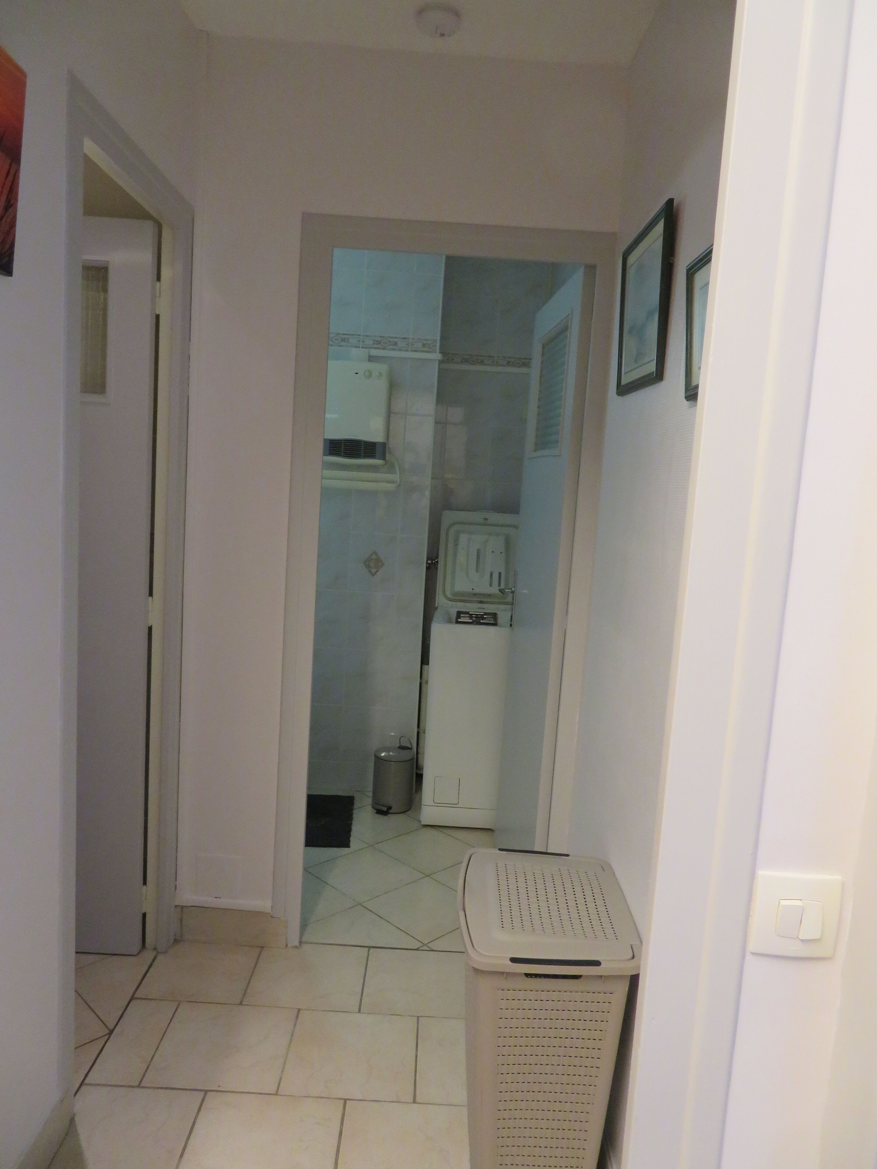 Een smalle gang met een wasmand, een kleine vuilnisbak en een wit apparaat, waarschijnlijk een koelkast, op de achtergrond.
