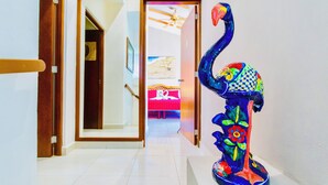 Un pasillo con una puerta de madera que da a una habitación con cama y ventilador de techo. Una escultura colorida se encuentra sobre el suelo.