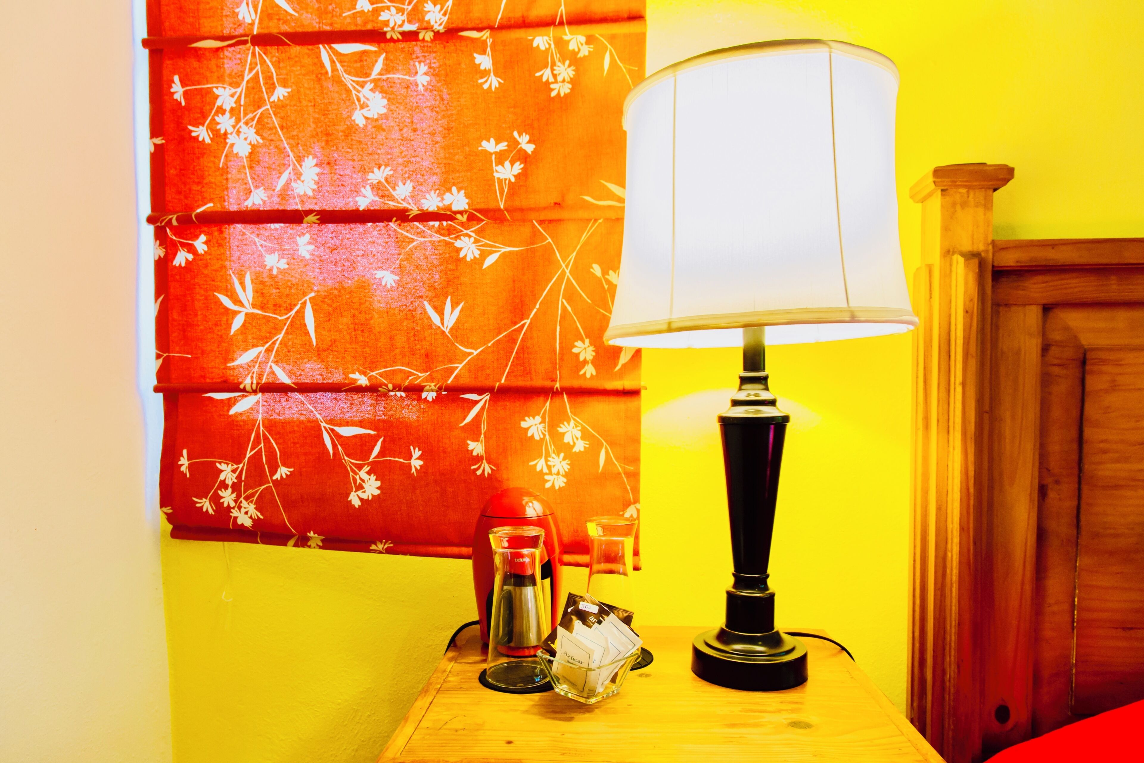Une pièce avec une table de chevet en bois, une lampe, un mur rouge et jaune, et un rideau à motifs floraux.