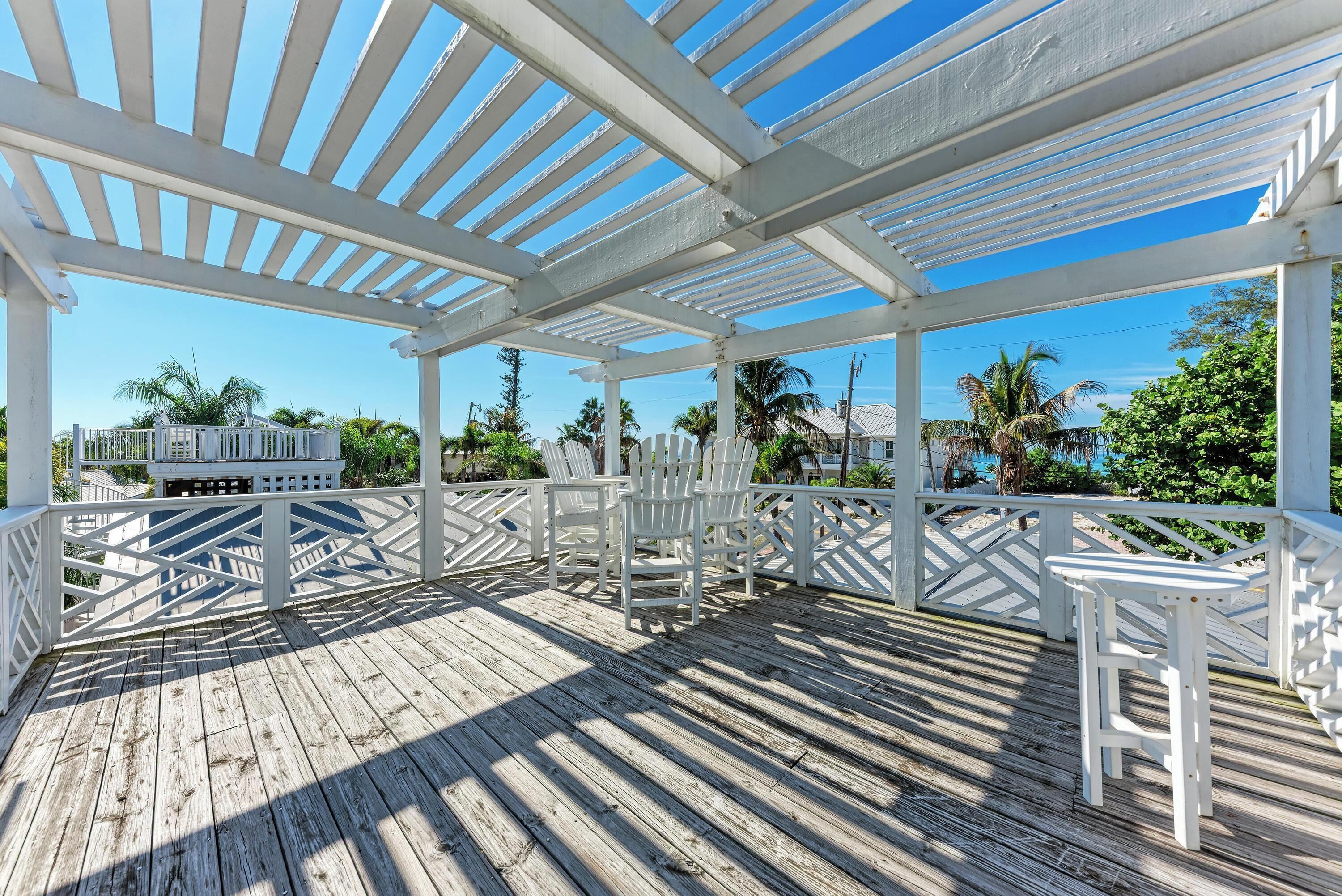 A covered wooden deck with white railings and chairs, overlooking a scenic view with palm trees and buildings.