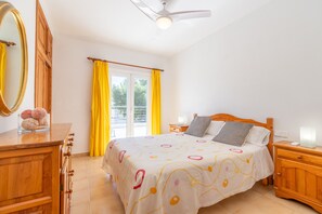 Een slaapkamer met een bed, houten nachtkastjes, een dressoir, een plafondventilator en een raam met gele gordijnen.