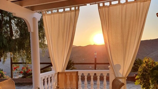A covered outdoor seating area with a table and chairs, a view of the sunset, and potted plants. - Peaceful Home Cortijo La Rijana with Pool, Terrific Views, Terrace, Wi-Fi (Sayalonga)