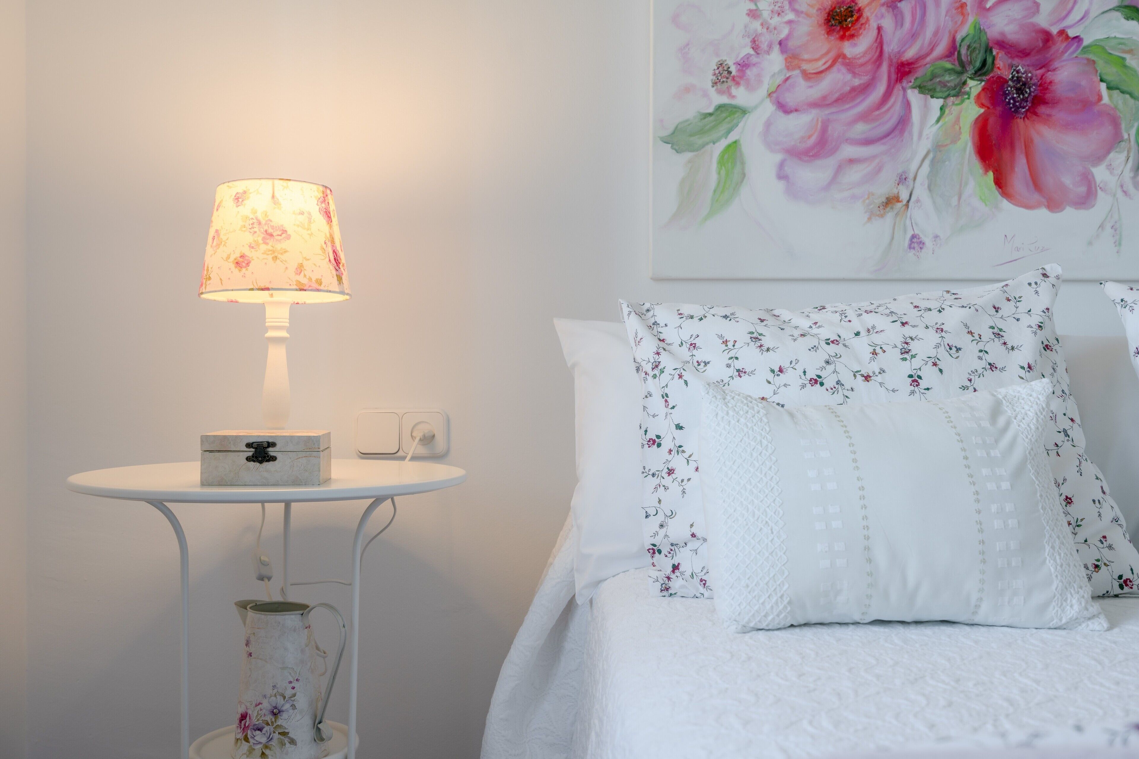 Une chambre à coucher avec un lit blanc, des coussins à motifs floraux, une table de chevet avec une lampe et un tableau floral accroché au mur.