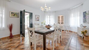 A dining area with a table set for six, white chairs, a chandelier, and a view through an open door. - Peaceful Home Cortijo La Rijana with Pool, Terrific Views, Terrace, Wi-Fi (Sayalonga)
