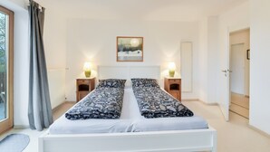 Una cama doble con colcha estampada, dos mesitas de noche con lámparas, un cuadro enmarcado en la pared y una ventana con cortinas.