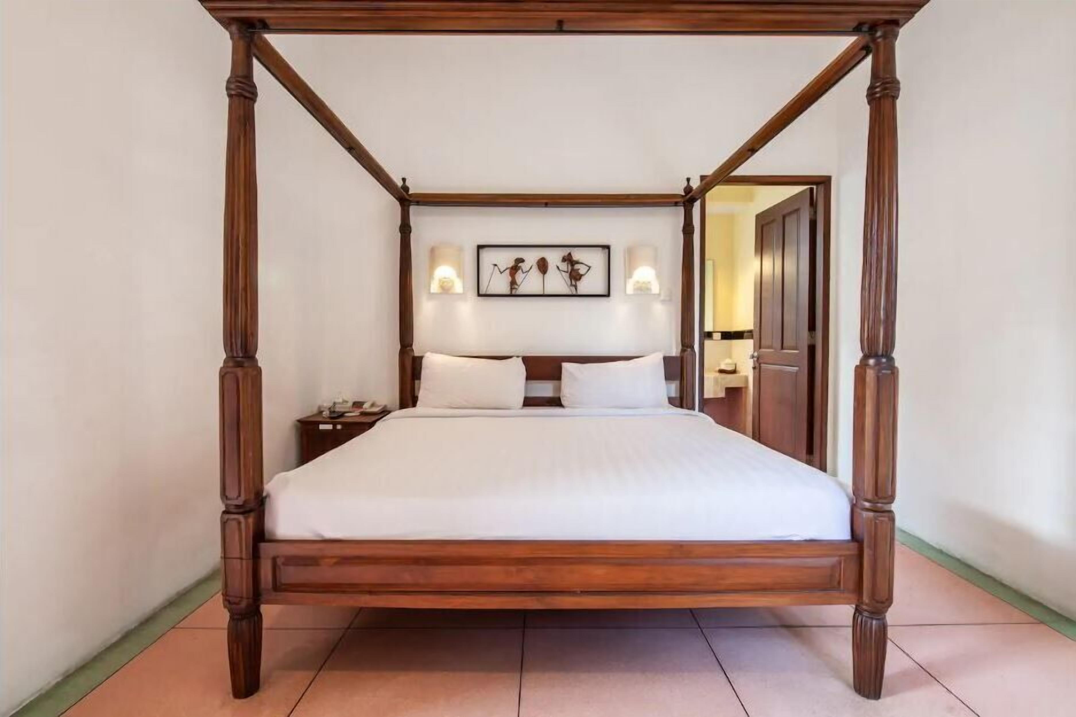 Una cama con dosel de estructura de madera, ropa de cama blanca y apliques de pared con una obra de arte encima.