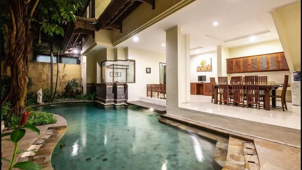 A swimming pool with a stone border, a wooden deck, and a dining area with chairs and a table. - PB - 7BR - V5 & V1 7BR Private Pool Villa, Walk to Seminyak Beach (Seminyak)